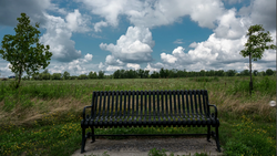 Black bench on a concrete pad facing an expansive green field under a partly cloudy sky.