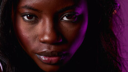 Close-up portrait of a woman with deep brown skin tone against a dark background with purple lighting.
