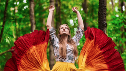 Woman with arms raised wearing flowing red and yellow fabric against green forest backdrop.