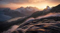 Snow-covered mountain peaks illuminated by golden sunrise light, with a glacial valley and frozen lake below.