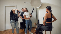 Photography studio setup with model posing on stool while crew adjusts octabox softbox lighting.