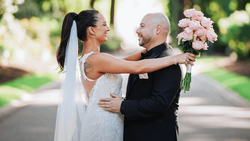 Bride and groom embrace outdoors while holding a bouquet of pink roses on their wedding day.