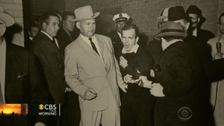 Men in suits and fedoras gathered in a brick corridor, one in a light tan coat being escorted by officials.