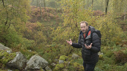 Man in black puffer jacket gesturing while speaking in a forested autumn setting.