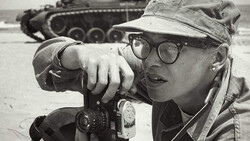 Photographer wearing glasses and cap looking through rangefinder camera with military tank blurred in background.