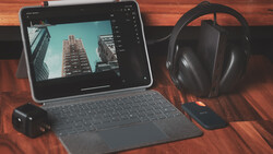 Tablet displaying architectural photography with editing software open, surrounded by headphones and external hard drive on wooden surface.