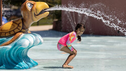 Child in pink swimwear crouching while being sprayed by a decorative turtle water feature.