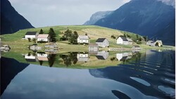 Alpine valley with scattered houses reflected in still lake water, surrounded by steep forested mountains.