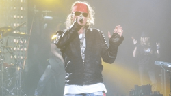 Performer on stage wearing sunglasses and leather jacket, singing into microphone with dramatic yellow stage lighting.