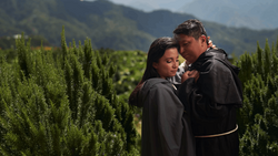 Two people in dark clothing embrace among tall green cypress trees with mountains visible in the soft-focused background.