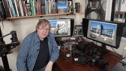 Photographer at desk with dual monitors displaying architectural images and camera equipment.