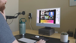 Podcaster or content creator speaking into a microphone at a desk setup with monitor displaying editing software.