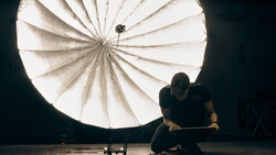 Photographer crouching beside a large parabolic reflector on a dark studio floor.