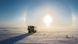 Military truck driving across a frozen landscape with sun dogs visible in the bright sky.