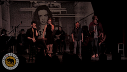 Band performing on stage with dramatic backlighting and a large portrait projection behind them.