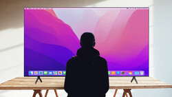 Person sitting at wooden desk viewing a large monitor displaying a vibrant pink and purple gradient wallpaper.