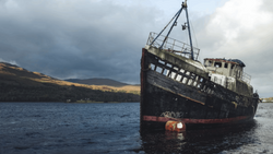 Abandoned ship with deteriorating hull tilted in shallow water against cloudy sky and barren hillside.