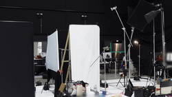 Professional photography studio setup with large white reflector panel, multiple light stands, and softbox lighting on black walls.