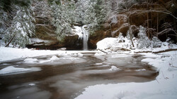 Frozen river with cascading waterfall framed by snow-covered evergreen trees and rocky cliffs.