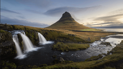 Twin waterfalls cascade into a dark river with a distinctive cone-shaped mountain rising behind moss-covered terrain.