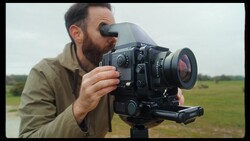 Cinematographer operating a film camera with viewfinder and attached lens in an open field.