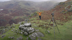 Surveyor with theodolite on tripod in mountainous valley landscape.