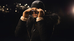 Person wearing dark winter jacket and sunglasses adjusting glasses against dark background with warm bokeh lights.
