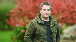 Man in olive jacket standing outdoors with red foliage in soft focus background.