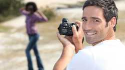Male photographer smiling at camera while holding a DSLR, with female subject posing in background on rural path.