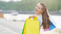 Woman holding colorful shopping bags at a waterfront location, smiling at camera.