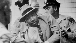 Black and white photograph of a man in a light-colored suit and fedora speaking with two police officers.