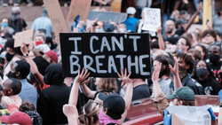 Protesters hold signs at a demonstration, including one reading "I CAN'T BREATHE" in white text on black.