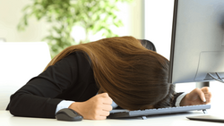 Woman in black blazer slumped over desk with head down, hand on keyboard, appearing exhausted or overwhelmed.
