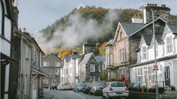 Misty mountains loom behind a quiet village street lined with parked cars and traditional stone buildings.