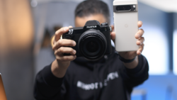 Person holding a black Fujifilm camera in one hand and a white smartphone in the other.