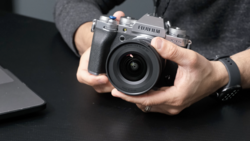 Hands holding a Fujifilm mirrorless camera with attached lens over a dark surface.