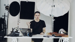 Photographer gesturing while seated at desk surrounded by professional camera equipment and lighting gear.