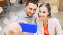 Two professionals smiling while taking a selfie with a smartphone in an office setting.
