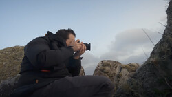 Photographer crouched on rocky terrain, holding a camera to their face while composing a shot against a cloudy sky.