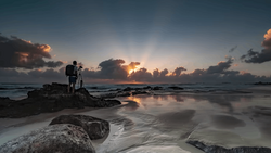 Photographer with tripod silhouetted on rocky outcrop during golden hour coastal sunset.