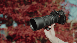 Photographer holding a mirrorless camera with a telephoto lens against a blurred red and turquoise floral background.