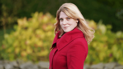 Woman with blonde hair wearing a red blazer, photographed outdoors against blurred green foliage.