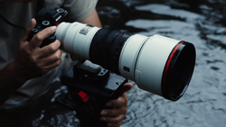 Photographer holding a mirrorless camera with a white telephoto lens featuring a red accent ring.