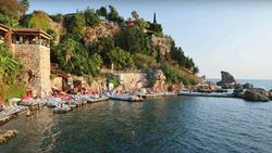 Mediterranean coastal settlement with stone buildings built into a hillside overlooking a calm bay with boats.