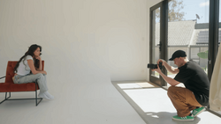 Photographer crouching while shooting a model seated on a red chair in a minimalist white studio.