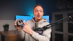 Man in podcast studio holding a vintage digital camera while speaking into a microphone.
