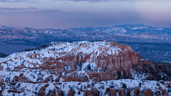 Snow-covered hoodoo formations and ridges in red rock canyon landscape with distant mountains under twilight sky.