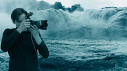 Photographer holding a DSLR camera while standing before massive ocean waves crashing with white spray.