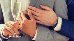 Two people wearing wedding rings hold champagne glasses while touching hands.