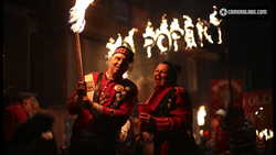 Two people in red ceremonial robes holding flaming torches during a nighttime procession.